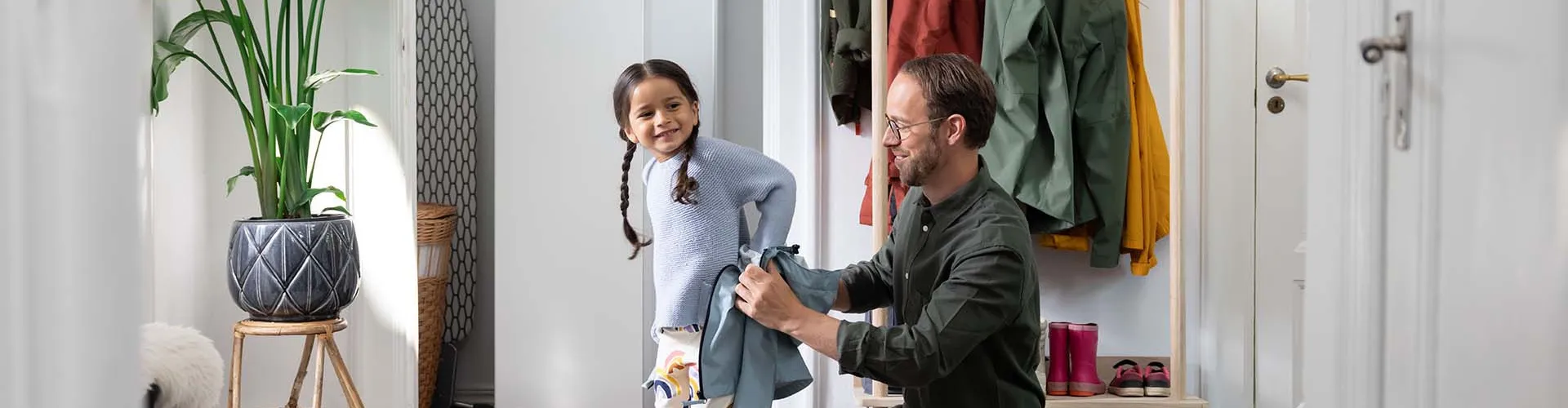 Ein Vater hilft seiner kleinen Tochter im Flur beim Anziehen, während im Hintergrund eine moderne Wärmepumpen-Inneneinheit von NIBE steht.