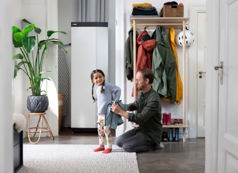 Ein Vater hilft seiner kleinen Tochter im Flur beim Anziehen, während im Hintergrund eine moderne Wärmepumpen-Inneneinheit von NIBE steht.