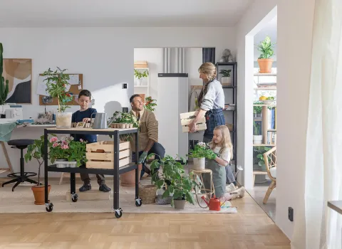 Eine vierköpfige Familie arbeitet gemeinsam in einem hellen Wohnzimmer an der Pflege von Zimmerpflanzen. Im Hintergrund steht eine moderne Wärmepumpen-Inneneinheit neben offenen Regalen und Pflanzenarrangements.