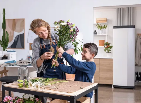Ein kleiner Junge hilft einer Frau beim Zusammenstellen eines frischen Blumenstraußes, während sie mit einer Schere die Stiele kürzt. Die Wärmepumpen-Inneneinheit von NIBE im Hintergrund passt zur gemütlichen Atmosphäre des hellen Raumes.