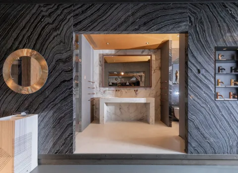 Ausstellungsbereich mit markanter Natursteinwand, rundem Spiegel, modernem Waschtisch aus weißem Marmor und Armaturen-Präsentation in der Badausstellung der Prehn & von Hoeßlin GmbH & Co. KG in Hamburg.