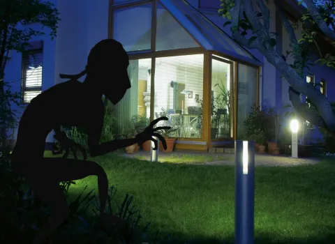 Ein beleuchteter Garten vor einem modernen Haus bei Nacht zeigt eine große Glasfassade mit Blick ins Wohnzimmer. Im Vordergrund ist die Silhouette eines Einbrechers zu sehen, der sich heimlich dem Haus nähert.