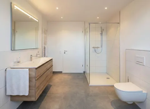 Fotorealistischer Planungsentwurf eines geräumigen Economy-Bades mit Glasdusche, Holzwaschtisch und großem Spiegel. Dezente Ausstattung für einen schlichten, eleganten Look.