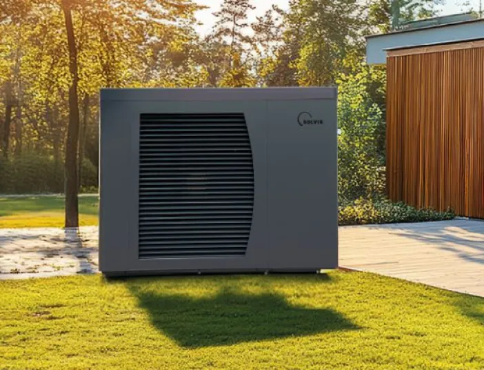 Eine Solvis-Wärmepumpe steht auf einer Rasenfläche vor einem modernen Mehrfamilienhaus mit Holzfassade, umgeben von Bäumen und warmem Sonnenlicht.