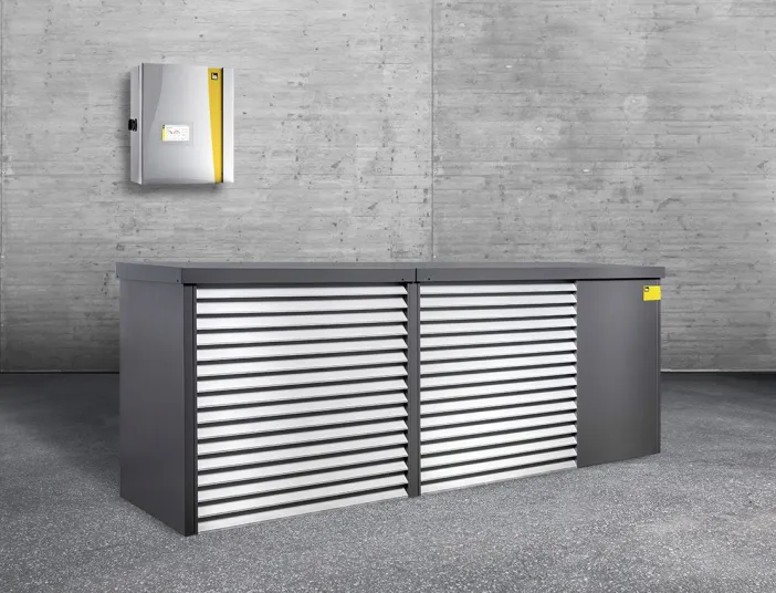 Eine Kaskadenlösung der intelligenten Luftwärmepumpe iDM Aero Alm Max steht in einem grauen Raum. Dahinter hängt die kleine Inneneinheit.